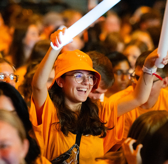 a smiling student holding large glow sticks in the middle of a sea of orange jams to a concert